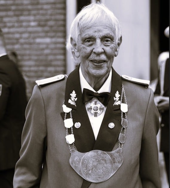 Older man in a formal military uniform with medals and ceremonial chains, standing outdoors at a ceremony.